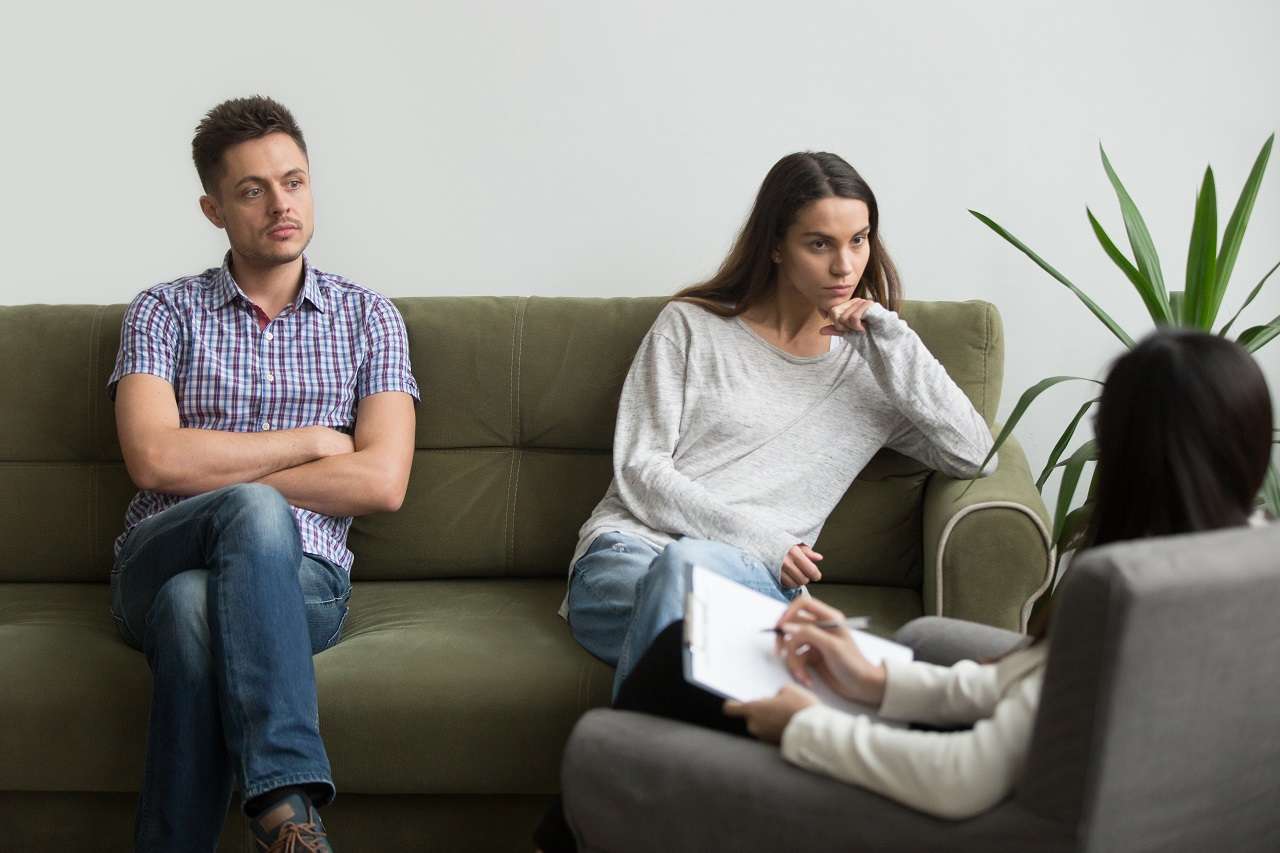 Unhappy millennial couple sitting apart on couch visiting female psychologist